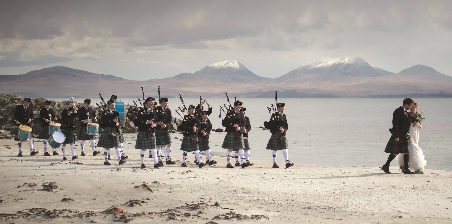 A couple that have just been married walk down the beach in Scotland with a traditional wedding pipe band